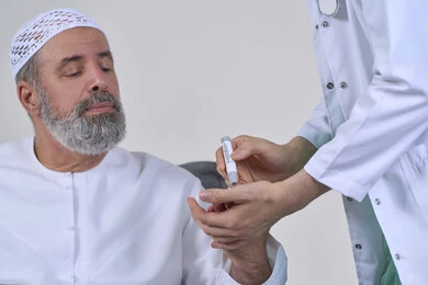 العمل في القطاع الطبي وشركات التمريض المنزلي، الرعاية الصحية والفحص الطبي السريع، بورتريه مقرب لممرض يرتدي المعطف الطبي يقوم بفحص نسبة السكر في الدم لمريض مسن عربي خليجي اماراتي يرتدي الكندورة يجلس على الكنبه، جهاز قياس سكر الدم لمرضى السكري، خلفية بيضاء