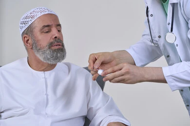 تقديم الرعاية الصحية والفحص الطبي السريع، جهاز قياس سكر الدم لمرضى السكري، العمل في القطاع الطبي وشركات التمريض المنزلي، بورتريه مقرب لممرض يرتدي المعطف الطبي يقوم بفحص نسبة السكر في الدم لمريض مسن عربي خليجي اماراتي يرتدي الكندورة يجلس على الكنبه، خلفية بيضاء