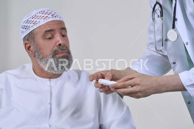 الفحص الطبي السريع والرعاية الصحية، جهاز قياس سكر الدم لمرضى السكري، العمل في القطاع الطبي وشركات التمريض المنزلي، بورتريه مقرب لممرض يرتدي المعطف الطبي يقوم بفحص نسبة السكر في الدم لمريض مسن عربي خليجي اماراتي يرتدي الكندورة يجلس على الكنبه، خلفية بيضاء