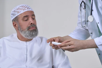 الفحص الطبي السريع والرعاية الصحية، جهاز قياس سكر الدم لمرضى السكري، العمل في القطاع الطبي وشركات التمريض المنزلي، بورتريه مقرب لممرض يرتدي المعطف الطبي يقوم بفحص نسبة السكر في الدم لمريض مسن عربي خليجي اماراتي يرتدي الكندورة يجلس على الكنبه، خلفية بيضاء