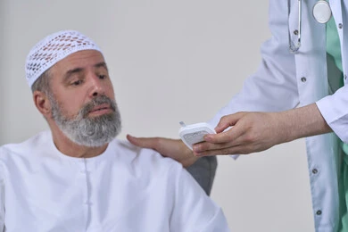 Healthcare and quick checkup, blood glucose meter for diabetics, working in the medical sector and home nursing companies, close-up portrait of a nurse in a medical coat checking the blood sugar level of an elderly Arab Gulf Emirati patient wearing a kandura sitting on a sofa, white background
