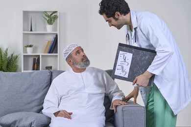 Working in home nursing companies, providing the best home medical services for elderly patients, following up on patients' medical records, portrait of a nurse wearing a uniform and a medical coat describing the medical condition of an elderly Arab Gulf Emirati patient wearing a kandura and sitting on a sofa, white background