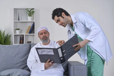 متابعة السجل الطبي للمرضى, العمل في شركات التمريض المنزلي، بورتريه لممرض يرتدي اليونيفورم والمعطف الطبي يقوم بوصف الحالة المرضية لمريض مسن عربي خليجي اماراتي يرتدي الكندورة ويجلس على الكنبه، تقديم أفضل الخدمات الطبية المنزلية لمرضى كبار السن، خلفية بيضاء