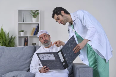 متابعة السجل الطبي للمرضى, العمل في شركات التمريض المنزلي، بورتريه لممرض يرتدي اليونيفورم والمعطف الطبي يقوم بوصف الحالة المرضية لمريض مسن عربي خليجي اماراتي يرتدي الكندورة ويجلس على الكنبه، تقديم أفضل الخدمات الطبية المنزلية لمرضى كبار السن، خلفية بيضاء