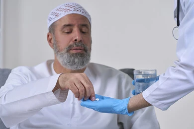 الاهتمام بالصحة والالتزام بالأوقات الصحيحة لأخذ العلاج, استخدام عقار الدواء بوصفة طبية, العمل في القطاع الطبي وشركات التمريض المنزلي، بورتريه مقرب لممرض يرتدي المعطف الطبي يقوم بإعطاء حبوب الدواء لمريض مسن عربي خليجي اماراتي يرتدي الكندورة، خلفية بيضاء