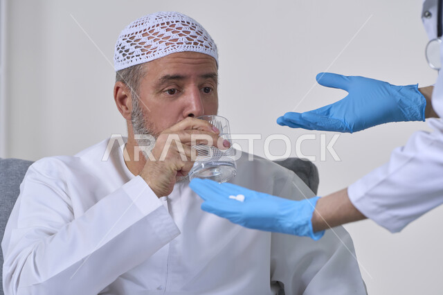 الاهتمام بالصحة والالتزام بالأوقات الصحيحة لأخذ العلاج, استخدام عقار الدواء بوصفة طبية, العمل في القطاع الطبي وشركات التمريض المنزلي، بورتريه مقرب لممرض يرتدي المعطف الطبي يقوم بإعطاء حبوب الدواء لمريض مسن عربي خليجي اماراتي يرتدي الكندورة، خلفية بيضاء