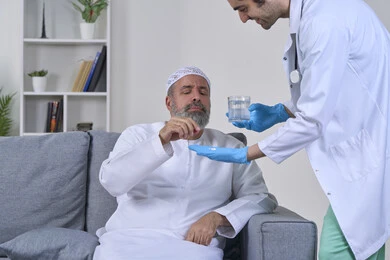 استخدام عقار الدواء بوصفة طبية, العمل في القطاع الطبي وشركات التمريض المنزلي، الاهتمام بالصحة والالتزام بالأوقات الصحيحة لأخذ العلاج, بورتريه لممرض يرتدي المعطف يقوم بإعطاء حبوب الدواء والماء لمريض مسن عربي خليجي اماراتي يرتدي الكندورة يجلس على كنبه، خلفية بيضاء