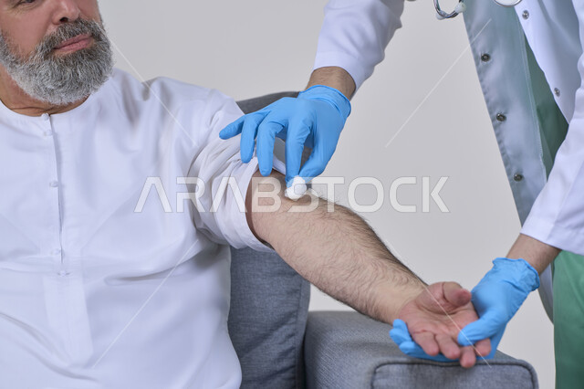 أخذ حقنة مسكنة للألم، الخدمات الطبية المنزلية للمرضى كبار السن، العمل في شركات التمريض المنزلي، بورتريه مقرب لممرض يرتدي اللباس الطبي يقوم يتعقيم مكان الابرة لرجل كبير بالسن عربي خليجي اماراتي يرتدي الكندورة ويجلس على كنبه، خلفية بيضاء