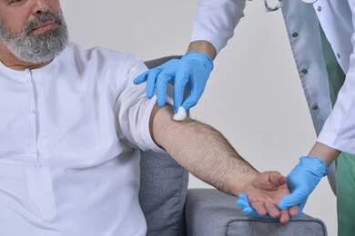 أخذ حقنة مسكنة للألم، الخدمات الطبية المنزلية للمرضى كبار السن، العمل في شركات التمريض المنزلي، بورتريه مقرب لممرض يرتدي اللباس الطبي يقوم يتعقيم مكان الابرة لرجل كبير بالسن عربي خليجي اماراتي يرتدي الكندورة ويجلس على كنبه، خلفية بيضاء