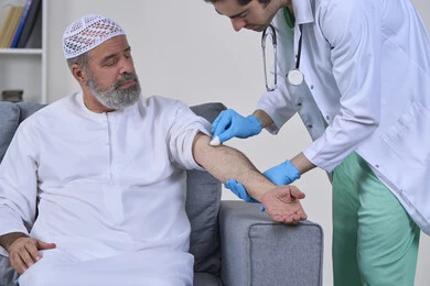 إعطاء حقنة مسكنة للألم، العمل في شركات التمريض المنزلي، تقديم أفضل الخدمات الطبية المنزلية للمرضى كبار السن، بورتريه لممرض يرتدي اللباس الطبي يقوم يتعقيم مكان الابرة لرجل كبير بالسن عربي خليجي اماراتي يرتدي الكندورة ويجلس على كنبه، خلفية بيضاء