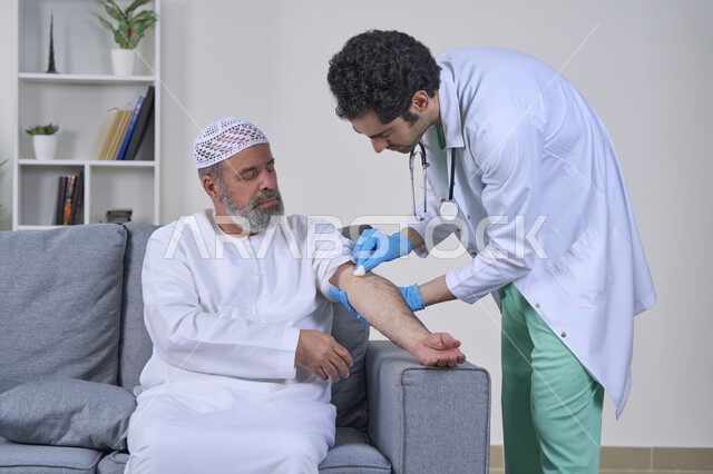 الخدمات الطبية المنزلية للمرضى كبار السن، أخذ حقنة مسكنة للألم، العمل في شركات التمريض المنزلي، بورتريه لممرض يرتدي اللباس الطبي يقوم يتعقيم مكان الابرة لرجل كبير بالسن عربي خليجي اماراتي يرتدي الكندورة ويجلس على كنبه، خلفية بيضاء