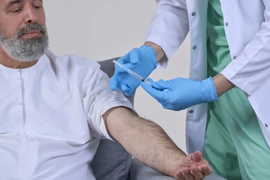 العمل في شركات التمريض المنزلي، أخذ الحقنة المسكنة للألم، تقديم أفضل الخدمات الطبية المنزلية للمرضى كبار السن، بورتريه مقرب لممرض اسعافات اولية يرتدي اللباس الطبي يقوم بإعطاء ابرة لرجل كبير بالسن عربي خليجي اماراتي يرتدي الكندورة ويجلس على كنبه، خلفية بيضاء