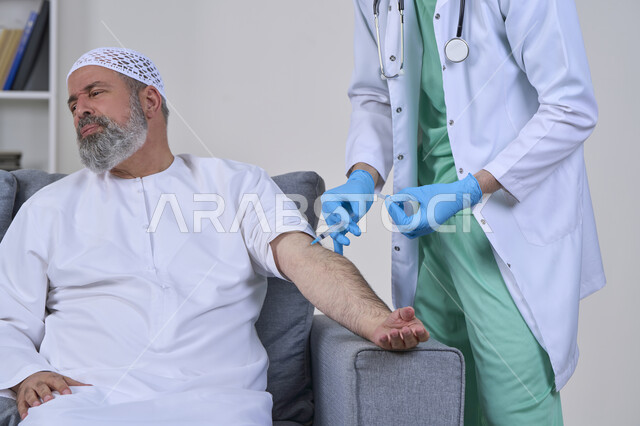 أخذ الحقنة المسكنة للوجع، تعابير الألم الشديد، تقديم أفضل الخدمات الطبية المنزلية للمرضى كبار السن، العمل في القطاع الصحي، بورتريه لممرض اسعافات اولية يرتدي اللباس الطبي يقوم بإعطاء ابرة لرجل كبير بالسن عربي خليجي اماراتي يرتدي الكندورة ويجلس على كنبه، خلفية بيضاء
