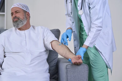 تعابير الألم الشديد، شركات التمرض المنزلي، تقديم أفضل الخدمات الطبية المنزلية للمرضى كبار السن، العمل في القطاع الصحي، بورتريه لممرض اسعافات اولية يرتدي اليونيفورم الطبي يقوم بإعطاء ابرة لرجل كبير بالسن عربي خليجي اماراتي يرتدي الكندورة ويجلس على كنبه، خلفية بيضاء