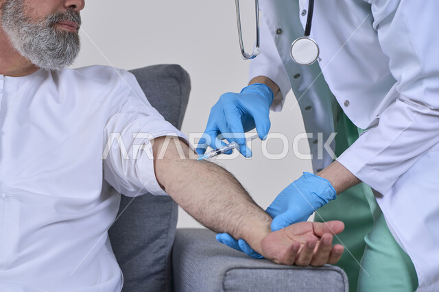 The concept of working in home nursing companies, taking a painkiller injection, providing the best home medical services for elderly patients, close-up portrait of a first aid nurse wearing medical clothing giving an injection to an elderly Arab Gulf Emirati man wearing a kandura and sitting on a sofa, white background