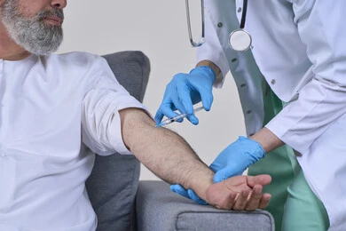 The concept of working in home nursing companies, taking a painkiller injection, providing the best home medical services for elderly patients, close-up portrait of a first aid nurse wearing medical clothing giving an injection to an elderly Arab Gulf Emirati man wearing a kandura and sitting on a sofa, white background