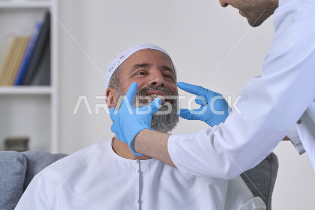 استخدام قشور فينير من الخزف لصنع ابتسامة بيضاء ناصعه، أهمية متابعة طبيب الاسنان، العناية والتجميل والتبيض، الفحص الطبي المنزلي بشكل دوري، بورتريه مقرب لممرض يرتدي القفازات الطبية يقوم بفحص أسنان مريض مسن عربي خليجي اماراتي يرتدي الكندورة والقبعة، خلفية بيضاء