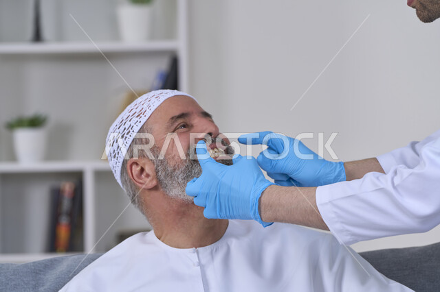 أهمية متابعة طبيب الاسنان، استخدام قشور فينير من الخزف لصنع ابتسامة بيضاء ناصعه، العناية والتجميل والتبيض، الفحص الطبي المنزلي بشكل دوري، بورتريه مقرب لممرض يرتدي القفازات الطبية يقوم بفحص أسنان مريض مسن عربي خليجي اماراتي يرتدي الكندورة والقبعة، خلفية بيضاء