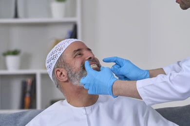 أهمية متابعة طبيب الاسنان، استخدام قشور فينير من الخزف لصنع ابتسامة بيضاء ناصعه، العناية والتجميل والتبيض، الفحص الطبي المنزلي بشكل دوري، بورتريه مقرب لممرض يرتدي القفازات الطبية يقوم بفحص أسنان مريض مسن عربي خليجي اماراتي يرتدي الكندورة والقبعة، خلفية بيضاء