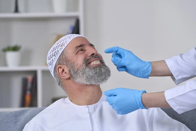 العناية والتجميل والتبيض، أهمية متابعة طبيب الاسنان، استخدام قشور فينير من الخزف لصنع ابتسامة بيضاء ناصعه، الفحص الطبي المنزلي بشكل دوري، بورتريه مقرب لممرض يرتدي القفازات الطبية يقوم بفحص أسنان مريض مسن عربي خليجي اماراتي يرتدي الكندورة والقبعة، خلفية بيضاء