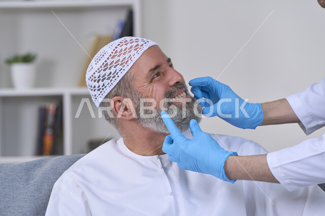 استخدام قشور فينير من الخزف لصنع ابتسامة بيضاء ناصعه، أهمية متابعة طبيب الاسنان، العناية والتجميل والتبيض، الفحص الطبي المنزلي بشكل دوري، بورتريه مقرب لممرض يرتدي القفازات الطبية يقوم بفحص أسنان مريض مسن عربي خليجي اماراتي يرتدي الكندورة والقبعة، خلفية بيضاء
