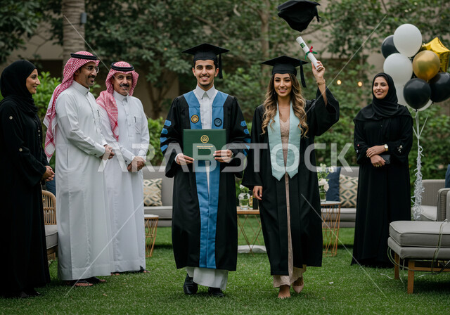 الاحتفال بإنهاء الدراسة الجامعية مع الاهل، مفهوم الاجتهاد والتفوق، طالبان عربيان سعوديان خليجيان يرتدي العباءه وقبعة التخرج ينظران إلى الكاميرا بإيماءات السرور والسعادة, الشعور بالفرح والنجاح