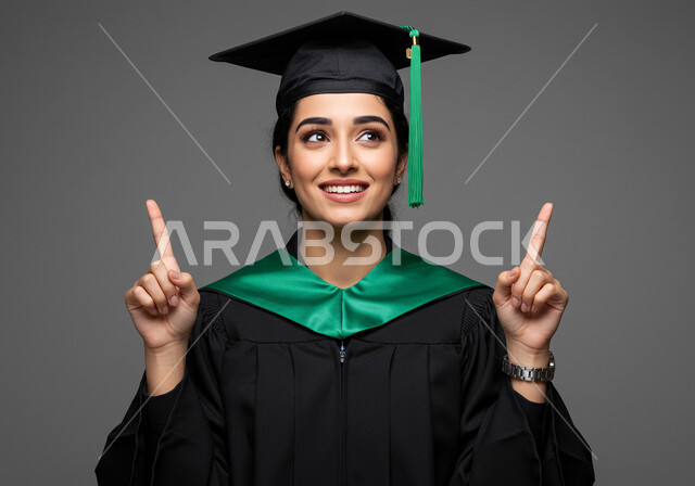 الشعور بالفخر والثقة بالنفس، مفهوم الاجتهاد والتفوق، الاحتفال بإنهاء الدراسة الجامعية، تعابير السرور والسعادة بالنجاح، بورتريه مقرب لطالبة عربية خليجية سعودية ترتدي عباءه وقبعة التخرج ترفع اصابع يدها بإيماءات النصر والسرور، خلفية رمادية