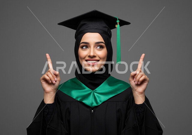 الشعور بالفخر والثقة بالنفس، مفهوم الاجتهاد والتفوق، الاحتفال بإنهاء الدراسة الجامعية، تعابير السرور والسعادة بالنجاح، بورتريه مقرب لطالبة عربية خليجية سعودية ترتدي عباءه وقبعة التخرج ترفع اصابع يدها بإيماءات النصر والسرور، خلفية رمادية