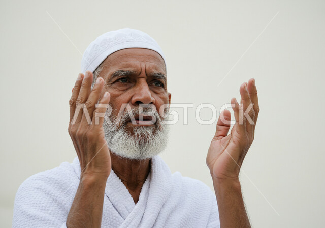 مفهوم أركان الإسلام وأداء الفرائض الدينية، رفع اليدين والدعاء بخشوع وتضرع، بورتريه مقرب لحاج خليجي عربي سعودي يرتدي ثياب الإحرام يقوم بتأدية مناسك الحج والعمرة، العبادة والتقرب إلى الله، خلفية بيضاء