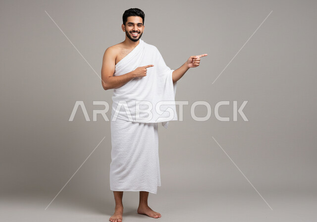 العبادة والتقرب إلى الله، مفهوم أركان الإسلام وأداء الفرائض الدينية، بورتريه لرجل حاج يرتدي ثياب الإحرام يقوم بتأدية مناسك الحج والعمرة يشير الى مكان ما بتعابير وايماءات السرور، الجسم بالطول الكامل، خلفية بيضاء