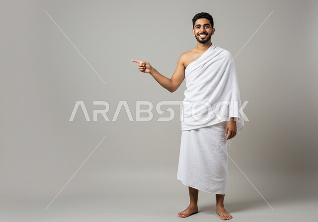 العبادة والتقرب إلى الله، مفهوم أركان الإسلام وأداء الفرائض الدينية، بورتريه لرجل حاج يرتدي ثياب الإحرام يقوم بتأدية مناسك الحج والعمرة يشير الى مكان ما بتعابير وايماءات السرور، الجسم بالطول الكامل، خلفية بيضاء