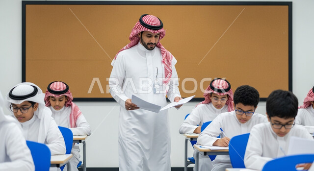 Education in the Kingdom of Saudi Arabia, studying and working hard for excellence, a Saudi Arabian Gulf Arab teacher wearing traditional dress monitors students during the end-of-semester exams, distributing exam papers