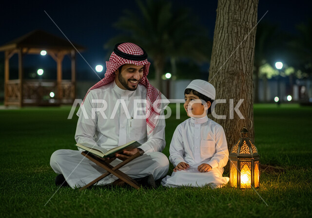 تلاوة وحفظ القرآن الكريم، زينة وفانوس شهر رمضان المبارك، جلسة خارجية في الهواء الطلق، تربية الاولاد على الفطرة الاسلامية الصحيحة، رجل عربي خليجي سعودي يرتدي الشماغ والثوب التقليدي يجلس على العشب يقرأ من المصحف الشريف بجانب ابنه في الليل