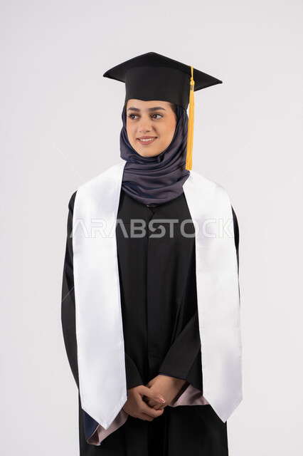 Standing up straight and taking selfies in graduation gown, portrait of a smiling Saudi Arabian Gulf woman wearing a graduation cap and gown looking at something with an expression of pleasure and happiness, Emirati graduate celebrating the completion of university, concept of excellence, success and diligence, white background