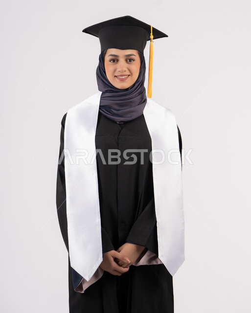An Emirati graduate celebrating the completion of university: Standing up straight and posing for selfies in her graduation gown. Portrait of a smiling Saudi Arabian Gulf woman wearing a graduation cap and gown, looking at the camera, expressing joy and happiness. Concept of excellence, success, and diligence. White background.