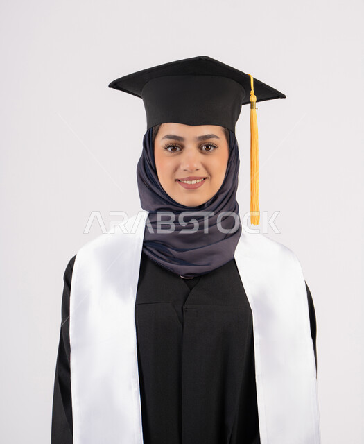 خريجة اماراتية تحتفل بإنهاء المرحلة الجامعية، الوقوف باستقامة والتقاط  صور شخصية بثياب التخرج، بورتريه مقرب لإمرأة عربية خليجية سعودية مبتسمة ترتدي القبعة وعباءة التخرج تنظر الى الكاميرا بتعابير السرور والسعادة، مفهوم التفوق والنجاح والاجتهاد، خلفية بيضاء