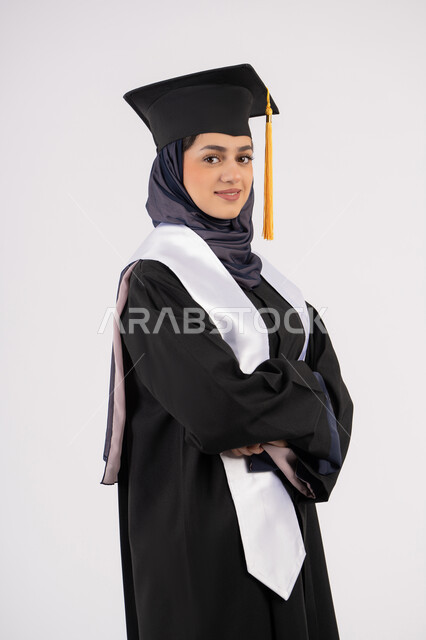 Celebrating the completion of university stage, an Emirati graduate standing with crossed hands and taking selfies in graduation gown, a portrait of a smiling Saudi Arabian Gulf woman wearing a graduation cap and gown with expressions of pleasure and self-confidence, the concept of excellence, success and diligence, white background