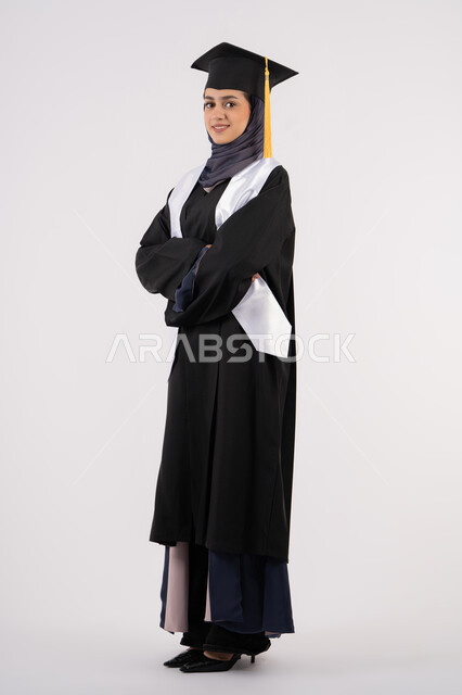 تعابير السرور والثقة بالنفس، خريجة اماراتية تقف بأيدي متقاطعة وتقوم بالتقاط  صور شخصية بثياب التخرج، بورتريه لإمرأة عربية خليجية سعودية مبتسمة ترتدي القبعة وعباءة التخرج تحتفل بإنهاء المرحلة الجامعية، التفوق والنجاح والاجتهاد، الجسم بالطول الكامل، خلفية بيضاء