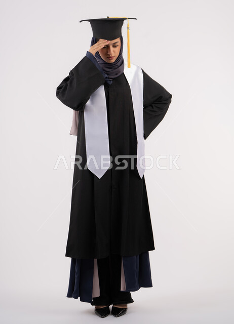 Fatigue and exhaustion after hard work and perseverance, portrait of a young Arab Gulf Emirati woman wearing a cap and graduation gown, putting her hand on her head with an expression of despair, finishing university studies after suffering, an Emirati graduate waiting for her results, full body, white background
