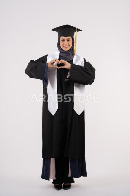 تشكيل اليد على شكل قلب،خريجة اماراتية تحتفل بإنهاء المرحلة الجامعية، بورتريه لشابة عربية خليجية سعودية مبتسمة ترتدي القبعة وعباءة التخرج تنظر الى الكاميرا بتعابير السرور والسعادة، مفهوم التفوق والنجاح والاجتهاد، الجسم بالطول الكامل، خلفية بيضاء