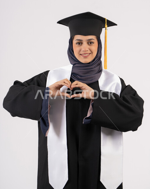 Hand forming a heart shape, Emirati graduate celebrating the completion of university, portrait of a smiling young Saudi Arabian Gulf woman wearing a graduation cap and gown looking at the camera with an expression of pleasure and happiness, the concept of excellence, success and diligence, white background