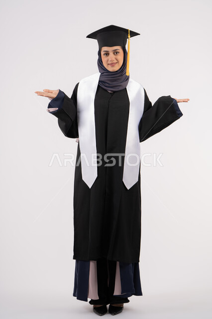 رفع اليدين لأعلى بتعابير الفرح، خريجة اماراتية تحتفل بإنهاء المرحلة الجامعية، بورتريه لشابة عربية خليجية سعودية مبتسمة ترتدي القبعة وعباءة التخرج تنظر الى الكاميرا بايماءات السرور والسعادة، مفهوم التفوق والنجاح والاجتهاد، الجسم بالطول الكامل، خلفية بيضاء
