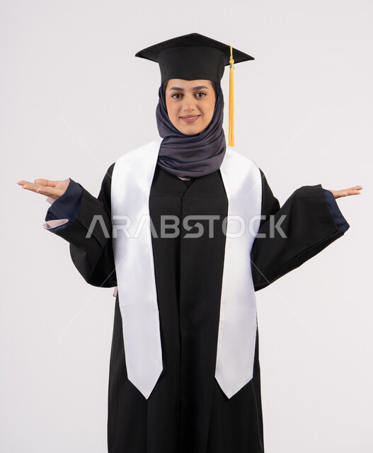 رفع اليدين لأعلى بتعابير الفرح، خريجة اماراتية تحتفل بإنهاء المرحلة الجامعية، بورتريه لشابة عربية خليجية سعودية مبتسمة ترتدي القبعة وعباءة التخرج تنظر الى الكاميرا بايماءات السرور والسعادة، مفهوم التفوق والنجاح والاجتهاد، خلفية بيضاء