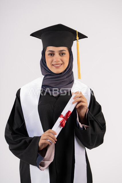 A young Saudi woman looking at the camera with gestures of pride, the concept of success and excellence, gestures of happiness at achieving a dream, a university occasion and celebration, celebrating the completion of university studies, a portrait of a veiled Arab Gulf Emirati graduate wearing a graduation gown and cap, holding a certificate in her hand, white background