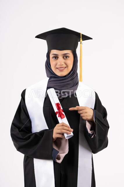 Hard work, success and excellence, a young Saudi woman looks at the camera with expressions of pride and happiness for achieving her dream, a university occasion and celebration, celebrating the completion of university studies, a portrait of a veiled Arab Gulf Emirati graduate wearing a graduation gown and cap, holding the certificate in her hand and pointing to it, white background