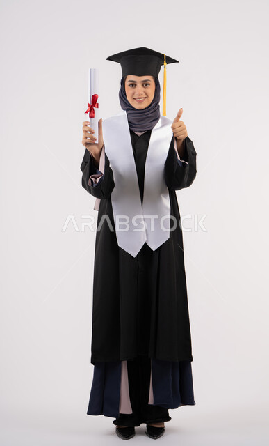 تعابير السعادة بالوصول الى الحلم، رفع اصبع الابهام بايماءات الرضى والاعجاب، مفهوم النجاح والتفوق، شابة سعودية تحتفل بإنهاء الدراسة الجامعية، مناسبة واحتفال جامعي، بورتريه لخريجة عربية خليجية اماراتية محجبة ترتدي زي التخرج والقبعة تحمل بيدها الشهادة، خلفية بيضاء