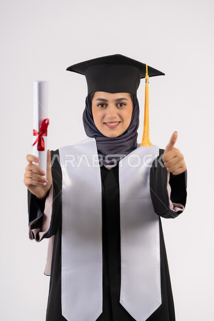 رفع اصبع الابهام بايماءات الرضى والاعجاب، تعابير السعادة بالوصول الى الحلم، مفهوم النجاح والتفوق، شابة سعودية تحتفل بإنهاء الدراسة الجامعية، مناسبة واحتفال جامعي، بورتريه لخريجة عربية خليجية اماراتية محجبة ترتدي زي التخرج والقبعة تحمل بيدها الشهادة، خلفية بيضاء