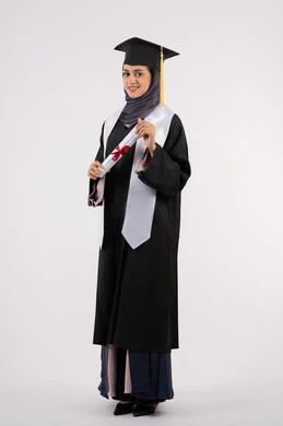A young Saudi woman looks at the camera with gestures of pride, expressions of happiness at achieving her dream, the concept of success and excellence, a university occasion and celebration, a celebration of completing university studies, a portrait of a veiled Arab Gulf Emirati graduate wearing a graduation gown and cap, holding a certificate in her hand, white background