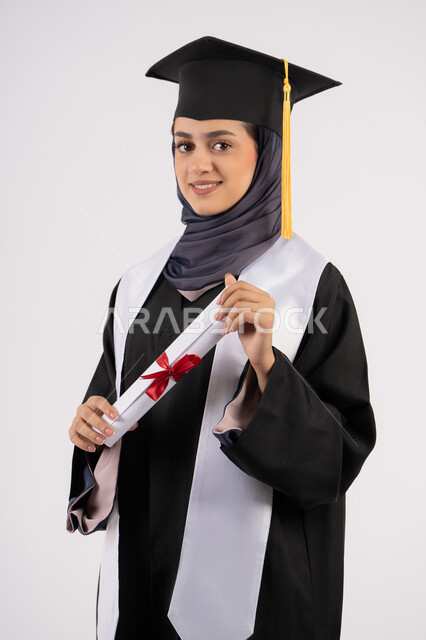 تعابير السعادة بالوصول الى الحلم، شابة سعودية تنظر الى الكاميرا بايماءات الفخر، مفهوم النجاح والتفوق، مناسبة واحتفال جامعي، الاحتفال بإنهاء الدراسة الجامعية، بورتريه لخريجة عربية خليجية اماراتية محجبة ترتدي زي التخرج والقبعة تحمل بيدها الشهادة، خلفية بيضاء