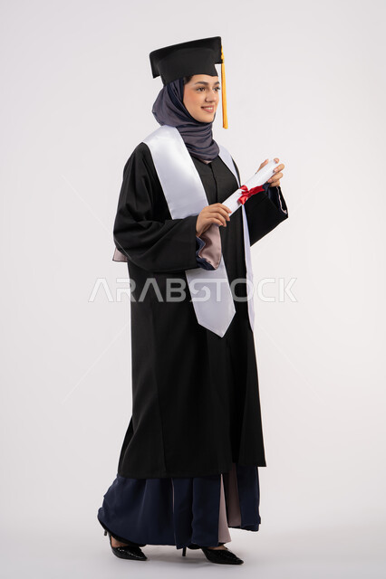 تعابير السعادة بالوصول الى الحلم، مفهوم النجاح والتفوق، الاحتفال بإنهاء الدراسة الجامعية، مناسبة واحتفال جامعي، شابة سعودية تنظر الى مكان ما بايماءات الفخر، بورتريه لخريجة عربية خليجية اماراتية محجبة ترتدي زي التخرج والقبعة تحمل بيدها الشهادة، خلفية بيضاء